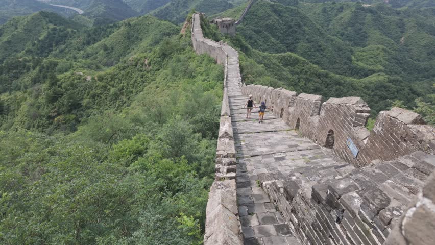A wide aerial shot showing people hiking along the ancient stone steps of the Great Wall of China, surrounded by lush green mountain ridges. Perfect for travel documentaries and heritage content.