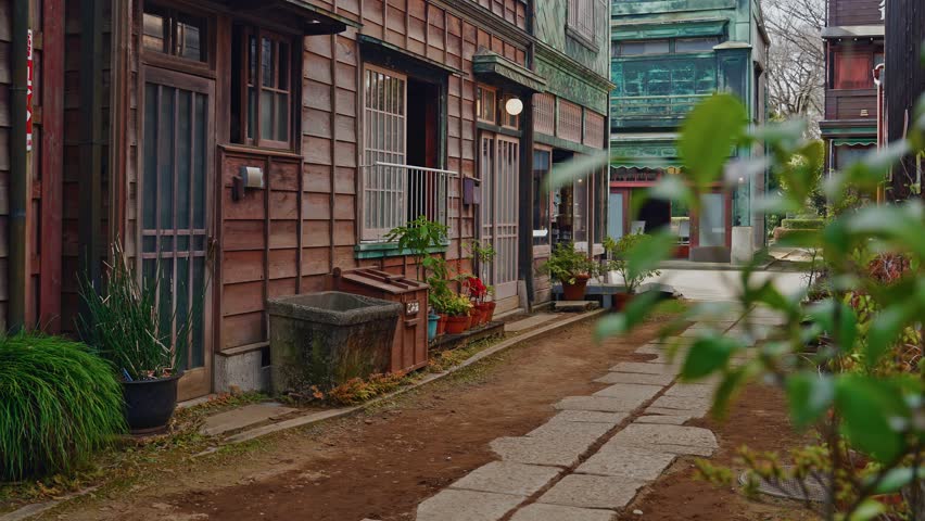 An establishing shot of a preserved Shitamachi street at Edo-Tokyo Open Air Architectural Museum, featuring weathered wooden facades, potted plants, and stone paths characteristic of the Meiji era.