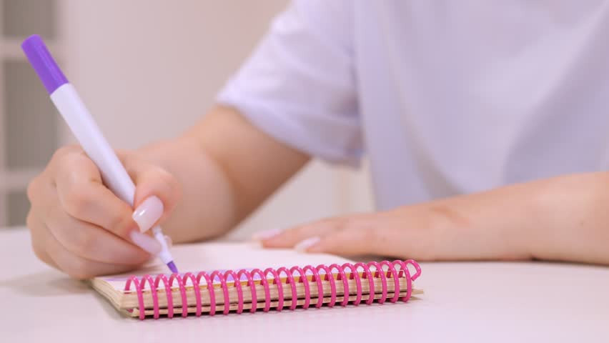 Hand writes with purple marker on pink spiral notebook. Soft focus highlights the pen and notebook. White shirt and neat fingernails add calm tone
