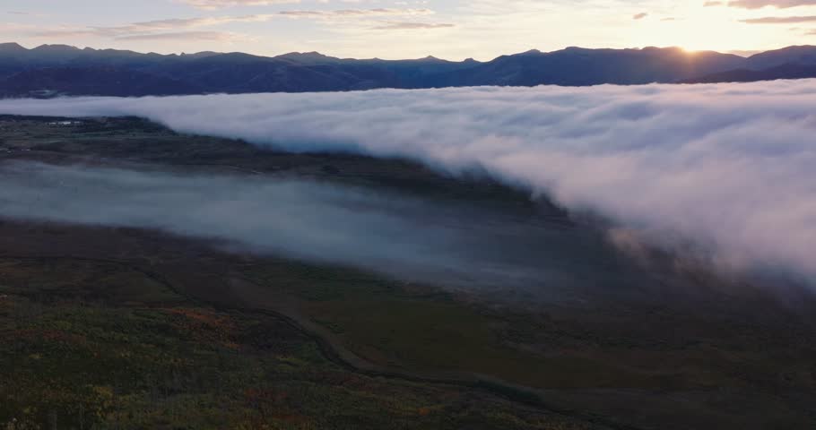 Aerial pullback above cloud inversion over Grand Lake Colorado, soft sunrise glow on mountain horizon, smooth drone