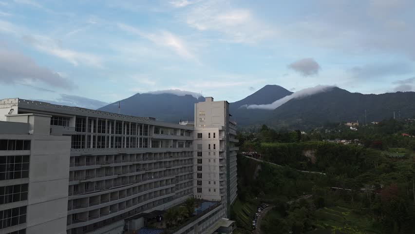 Panoramic aerial view of a city nestled at the foot of twin mountains under a soft blue evening sky. Low clouds drift across the mountain slopes, creating a dramatic and serene atmosphere. Urban buildings, residential houses, and lush green trees spread across the valley, blending natural landscape with city life in a tropical highland setting.