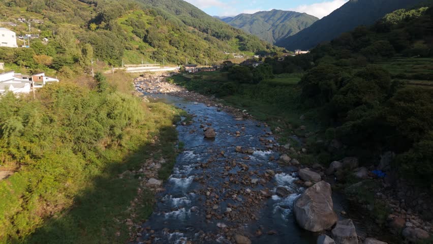 An aerial perspective captures a shallow, rocky river winding through a vibrant green valley between rolling forested mountains. Small houses and a quiet road line the riverbank, with the foreground showing the clear rippling water and the background featuring deep mountain ridges under a bright sky. Peaceful atmosphere of natural harmony