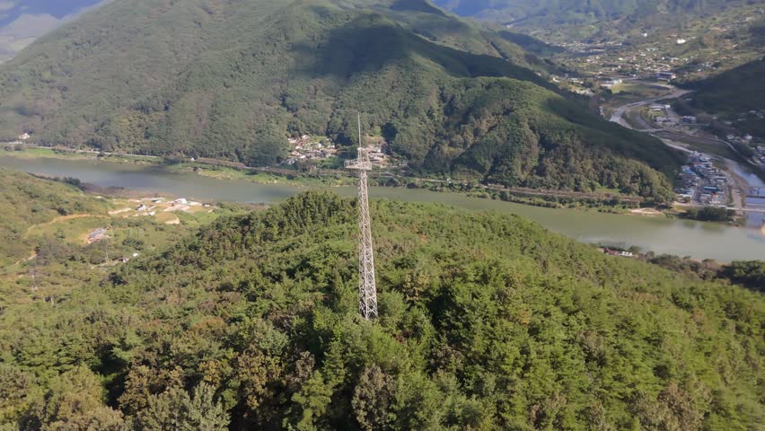 A tall silver lattice communication tower stands prominently amidst a dense green forest on a mountainside. In the background, a wide river flows through a valley surrounded by rolling hills and a small rural settlement under a clear sky. Concept of remote connectivity and infrastructure.
