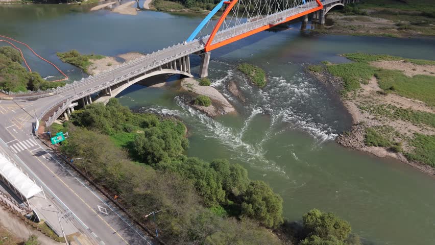 An aerial perspective captures a modern orange and blue arched bridge spanning a wide green river with rushing white water rapids. Lush green willow trees and dense vegetation line the rocky banks against a background of hills and clear sky. Concept of engineering and nature harmony.