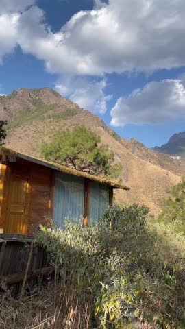 A cinematic 1080p aerial time-lapse capturing fast-moving white clouds over a rustic wooden cabin nestled against a majestic mountain range in Western China.