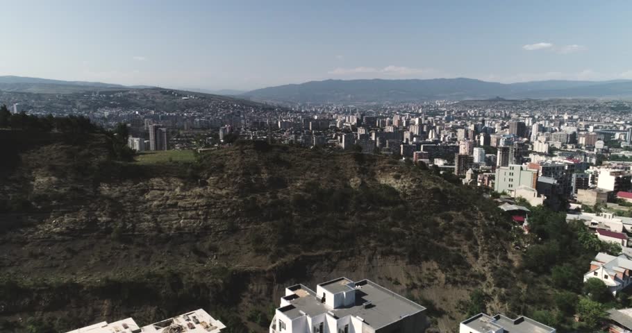 Wide drone aerial view of a large urban cityscape with high-rise buildings behind a rocky hillside, with a mountain range on the horizon under a clear blue sky. No people visible.
