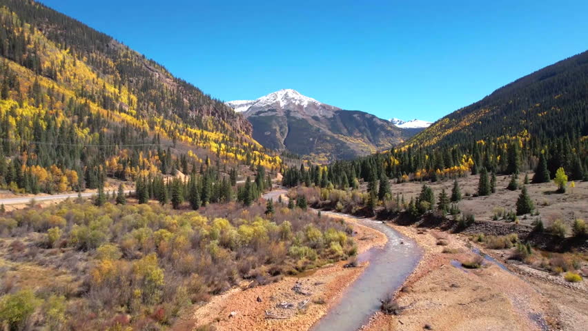 An aerial shot captures a vibrant autumn landscape featuring a winding road, lush green trees, and mountains in the background. The scene showcases the beauty of nature during fall with a clear blue sky.