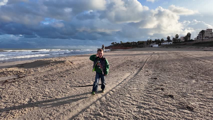 Little boy playing with stick on sandy beach near ocean waves under dramatic cloudy sky family vacation leisure outdoor childhood moment by the sea.