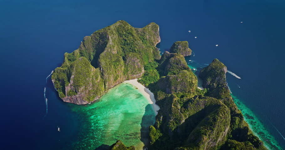 Phi Phi Island Maya Bay aerial: towering limestone cliffs draped in lush green, pristine white sand and vibrant turquoise water with boats cruising coast. Summer vacation paradise, travel to Thailand
