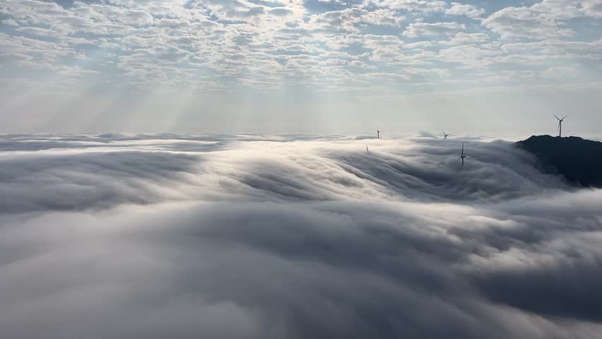 A cinematic 1080p aerial time-lapse capturing white wind turbines spinning atop a mountain peak, surrounded by a vast, fast-moving sea of clouds under a misty sky, China.