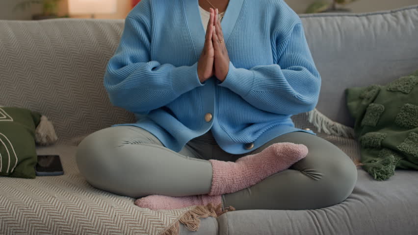 Tilt up shot of focused African American young lady practicing breathing exercise in lotus pose on couch in modern living room