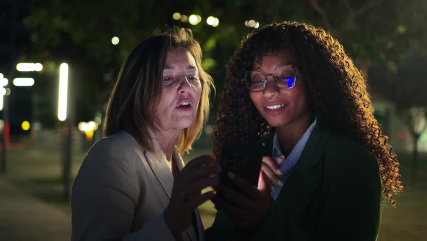 Two focused women collaborating on a smartphone, showcasing teamwork and communication in a lively nighttime atmosphere, symbolizing connection and innovation in a professional landscape.