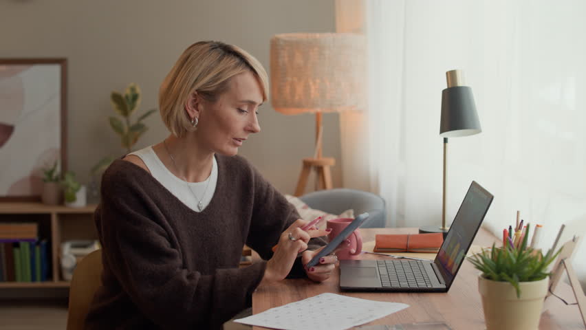 Medium shot of cheerful Caucasian businesswoman holding smartphone taking notes in paper calendar while sitting at table with wireless laptop, working from home