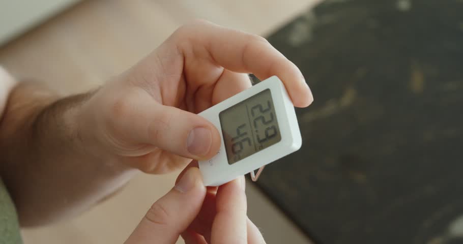 Close-up of a hand holding a digital thermometer and hygrometer, checking indoor temperature and humidity levels to prevent mold growth and maintain a healthy home environment.