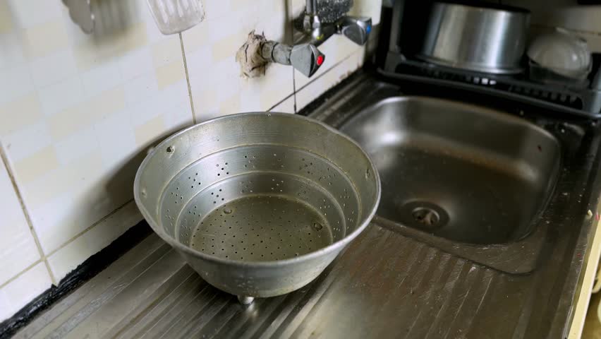 Old perforated metal colander placed on a stainless steel drainboard next to a kitchen sink with wall mounted faucet and stained tiled walls in natural indoor lighting.