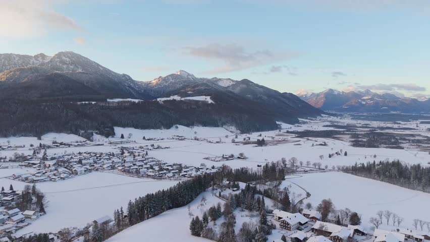 Aerial view of a snow covered alpine village at sunset with mountains in the background. Peaceful winter landscape with residential houses, church and rural countryside in Bavaria, Germany.