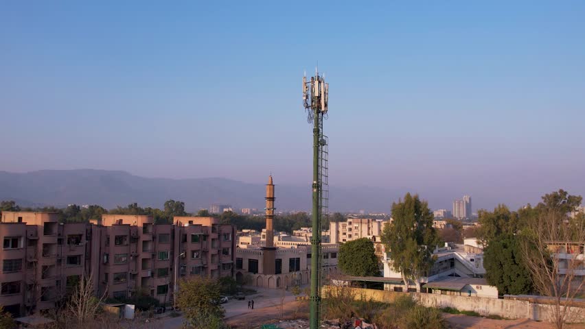 Elevated shot of a tall cell tower above residential buildings with distant hills under clear sky, Islamabad, Pakistan
