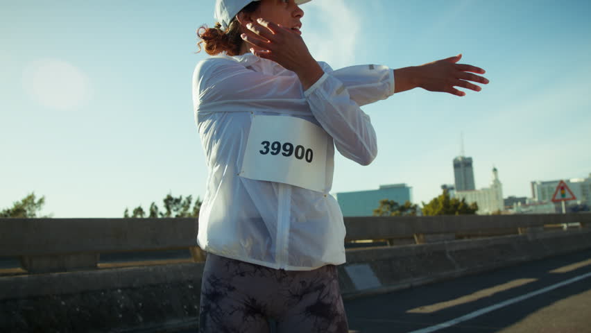 A focused female athlete stretches and warms up her muscles before participating in an urban marathon, preparing mentally and physically for the challenge ahead under a clear sky.