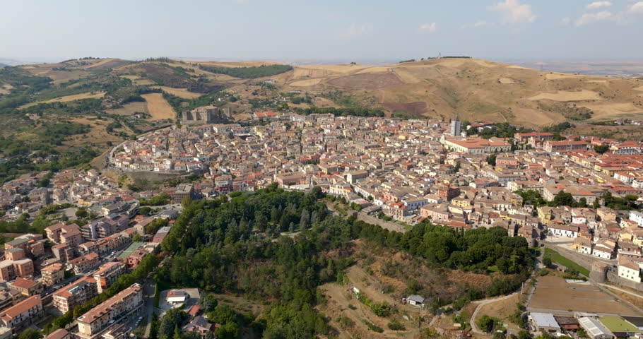 Aerial panorama of Melfi, a town in Basilicata, Southern Italy. It is an old Italian city with red-tiled roofs, nestled among rolling golden hills. A clear blue sky meets the horizon in the background
