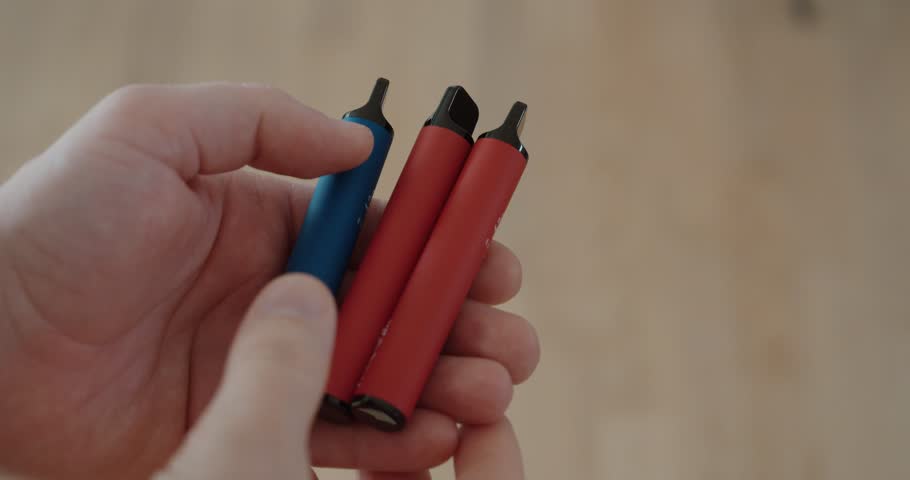 Close-up of a hand holding several disposable e-cigarettes, highlighting health risks, nicotine consumption, and environmental concerns related to electronic waste and recycling.