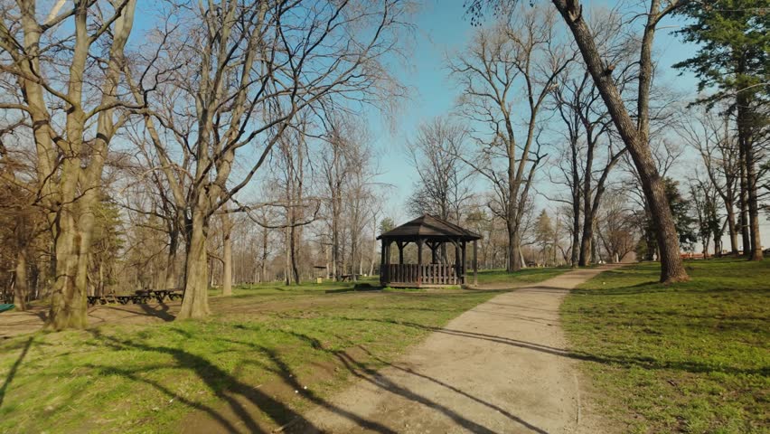 Wooden gazebo in a quiet park surrounded by leafless trees on a sunny early spring day. Concept of outdoor recreation, relaxation in nature and peaceful urban landscape