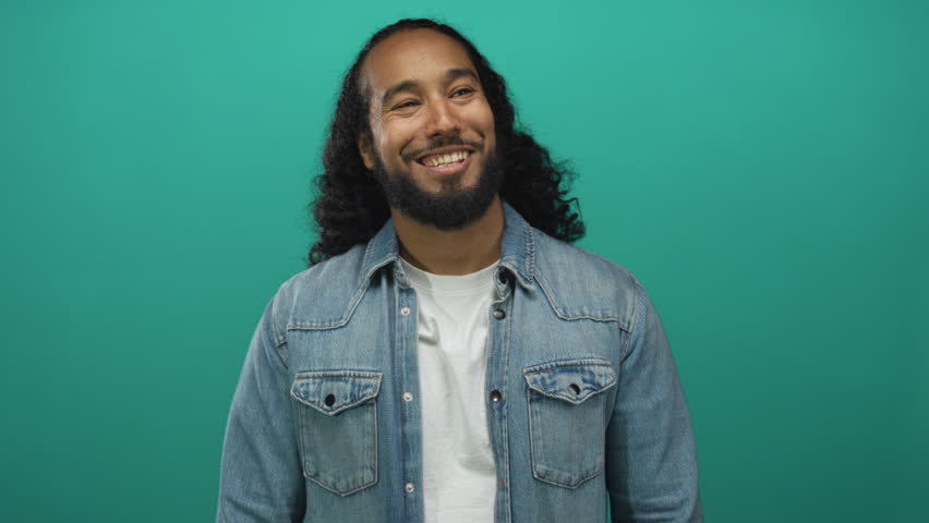 Man smiling with beard and teeth visible in studio against teal wall and denim jacket; joy confidence relaxed.