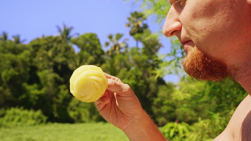 Man Eating Whole Peeled Mini Pineapple Outdoors Surrounded by Tropical Green Nature Under Bright Sunlight, Close Up View Showing Exotic Fruit Snack and Summer Lifestyle.