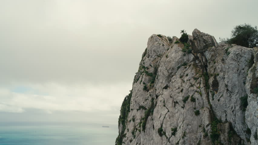 Dramatic view of the famous Rock of Gibraltar towering above the Mediterranean Sea under cloudy skies, with distant ships on the horizon.