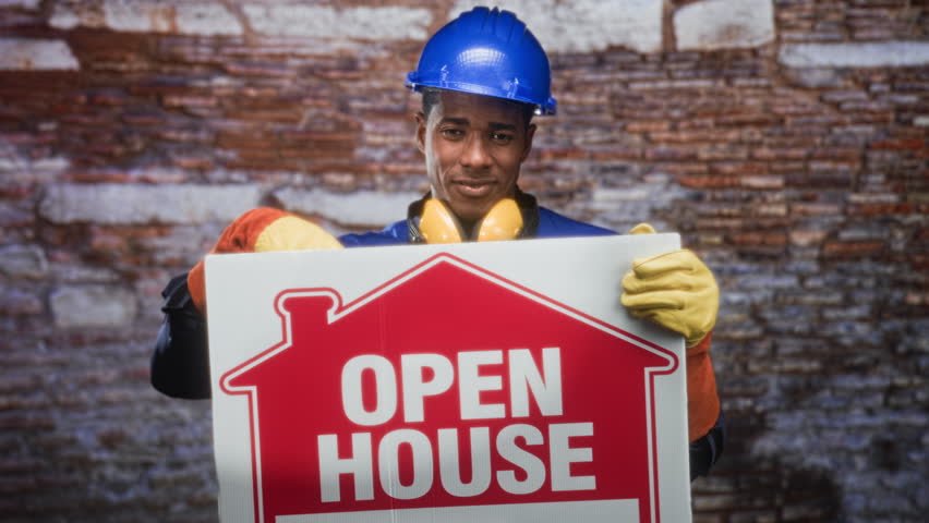 Man builder holding an open house sign and pointing finger to temple in front of a brick building wall; confidence invitation.