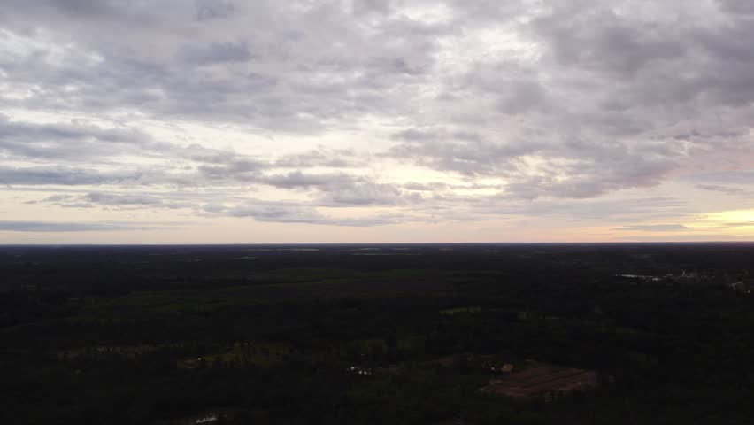 Sideward flying circling view of sunset sky over rural tropical landscape with sporatic building along the land and cloudy overcast sky.