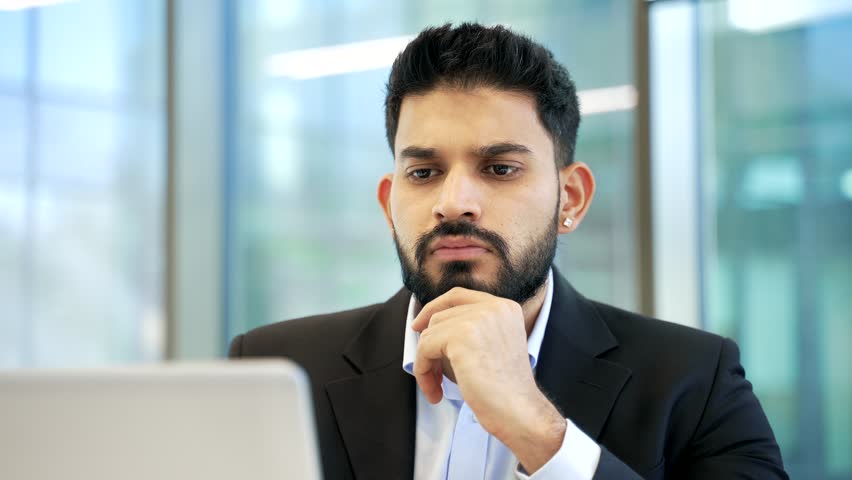 Businessman thinking about problem solving while working on laptop sitting at workplace in business office. Thoughtful serious manager in formal suit is busy with startup project on computer. Close up
