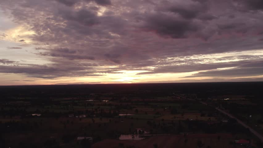 High altitude panning motion flying motion view of the setting sun over rural rtropical landcape, with couldy sky and hazy horizon in the background.