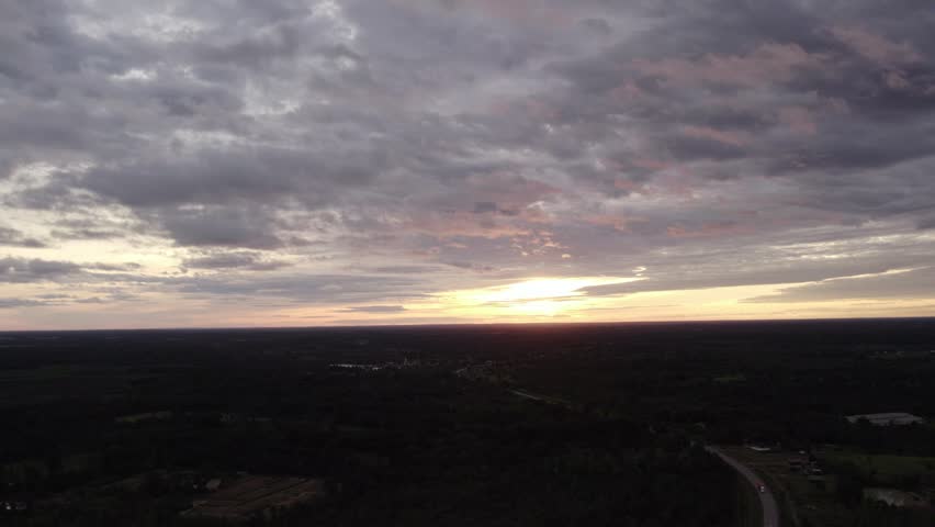 Panning dideward motion flying view of the setting sun over rural tropical landscape with sporatic building along the land and cloudy overcast sky.