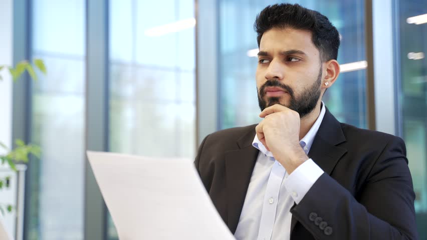 Serious businessman reviewing financial documents while sitting in modern business office. Thoughtful executive checking analyzing reports, contracts, investment data or corporate accounting papers. 