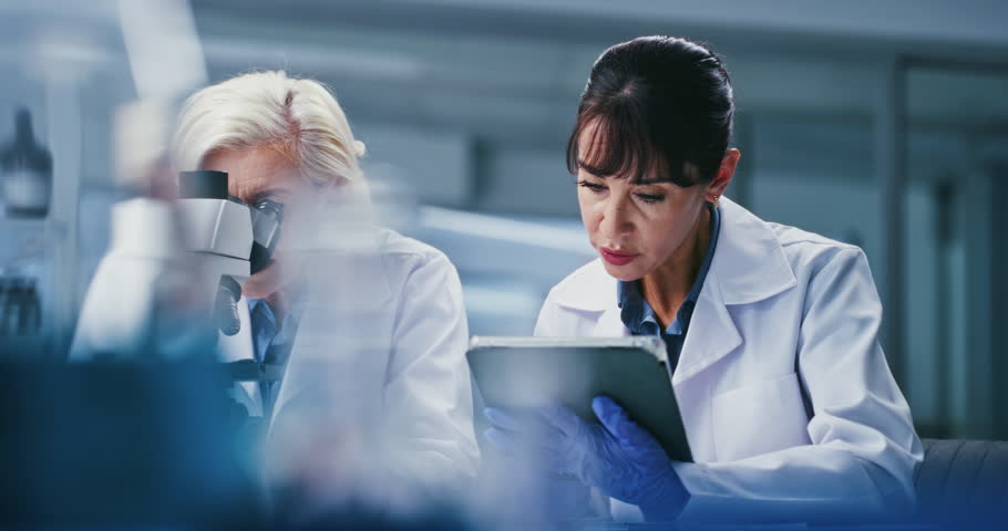 Microscope, talking and women on tablet in lab for teamwork, medical study and experiment results. Healthcare, mentor and scientists on tech for clinical trial, stem cell research and medicine review