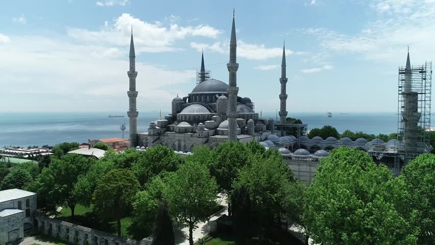 An aerial shot showcasing the majestic Blue Mosque in Istanbul, Turkey, with its iconic domes and minarets, surrounded by lush green trees and the sea in the background under a bright sky.
