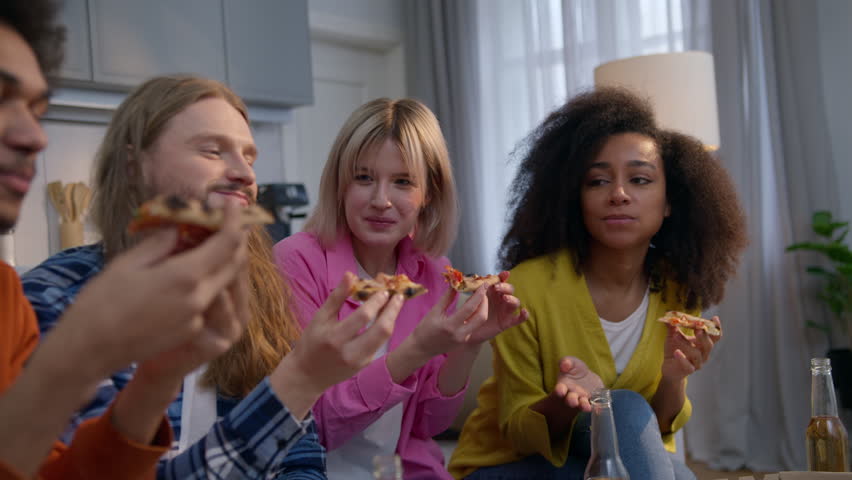 Happy young ethnic friends men and women eating pizza at home party couch sofa students enjoy meal together hungry Caucasian African girls guys drink beer alcohol talking multiethnic friendship indoor