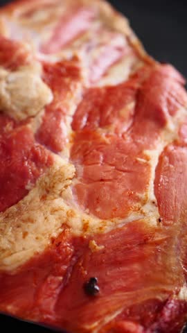 Raw cured pork, a hearty cut of meat displaying rich red muscle and white fat, rests on a dark, speckled ceramic plate.