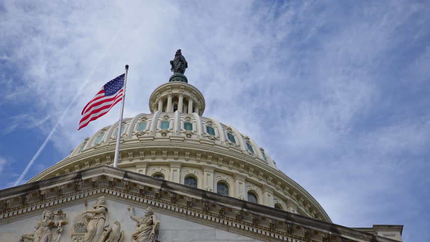 American flag waving on US Capitol dome. American flag in clouds over Capitol landmark. American flag symbol of Amerika democracy. American flag on historic Capitol in Washington DC.