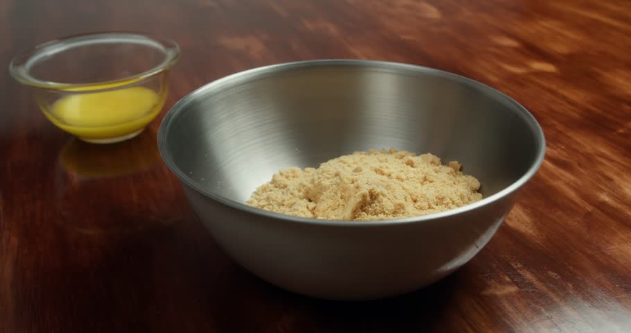 Crushed cookies mixed with melted butter in metal bowl to prepare cheesecake crust. Close up of homemade dessert base preparation on wooden table in kitchen.
