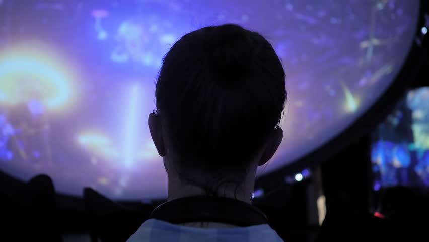Back view: woman wearing wireless black headphones and looking around in dark room of interactive exhibition or museum with colorful illumination. Futuristic, entertainment, immersive concept