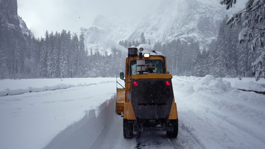 Yellow plow clearing mountain road in heavy snow, park maintenance operator drives across icy pathway flanked by towering granite cliffs and dense evergreen forest. Yosemite National Park