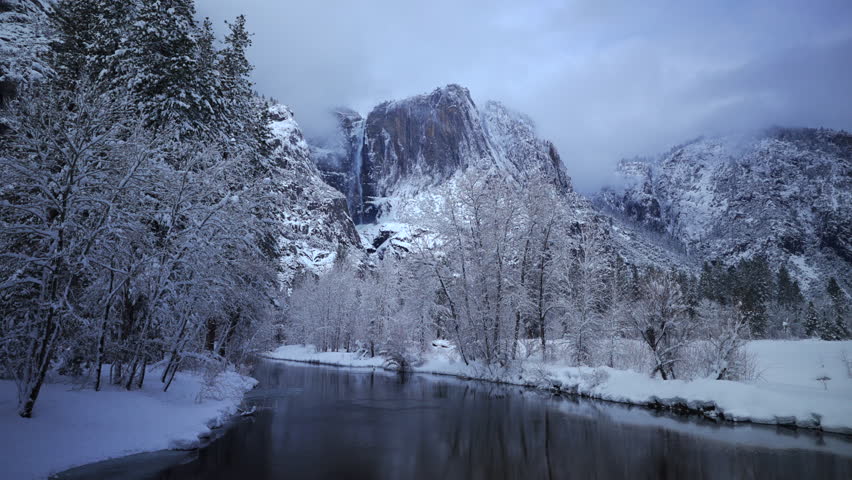 River reflection snowy cliffs in Yosemite National Park, tranquil blue hour scene with mirrorlike water and frozen banks, distant waterfall and towering granite illuminated by soft evening light,
