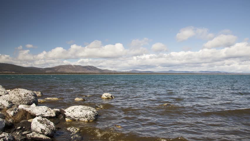 Eagle Lake in Lassen County, California captures a full hour of dynamic weather compressed into a single time lapse sequence. Cumulus clouds build and shift across a blue sky as strong winds drive visible chop across the surface, creating rhythmic wave patterns along the rocky shoreline. The foreground features scattered granite and basalt boulders partially submerged at the shore