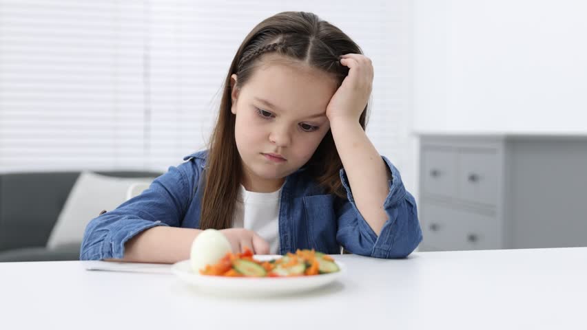 Unhappy little girl refusing to eat egg and vegetables at white table in kitchen