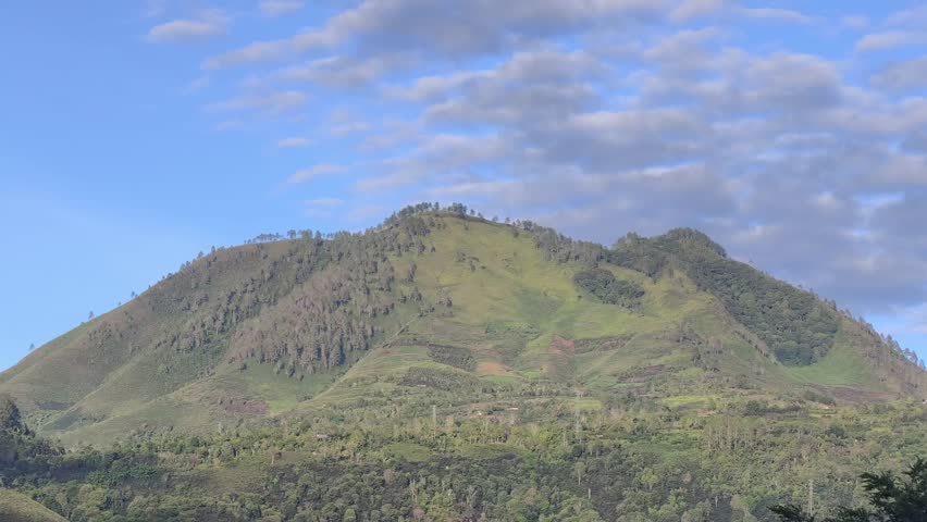 Scenic landscape of lush green hills and tropical mountains under a clear blue sky with white clouds.