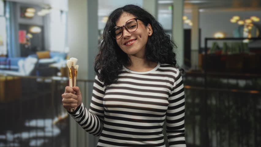 Woman holding makeup brushes and smiling in a hotel building lobby; beauty creativity confidence empowerment.