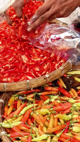 Close up of hands packing freshly sliced red chili peppers into plastic bags above baskets of colorful chili varieties at a busy traditional market.