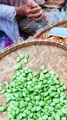 A close-up of a hand sorting green petai beans on a woven tray, surrounded by chili peppers and goods, highlighting traditional market food preparation.