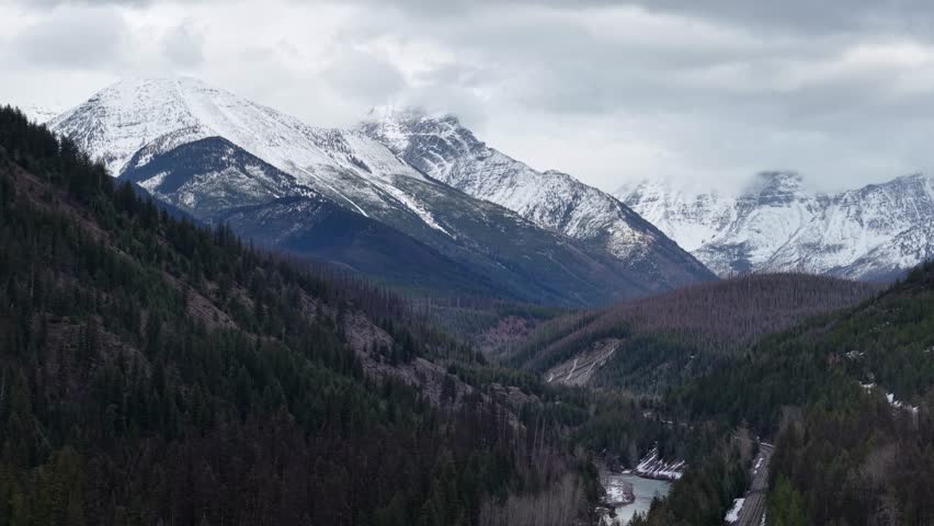 Drone aerial view of mountainous valley with winding road alongside pine forest and snow-covered peaks under cloudy sky in Glacier National Park, Montana.